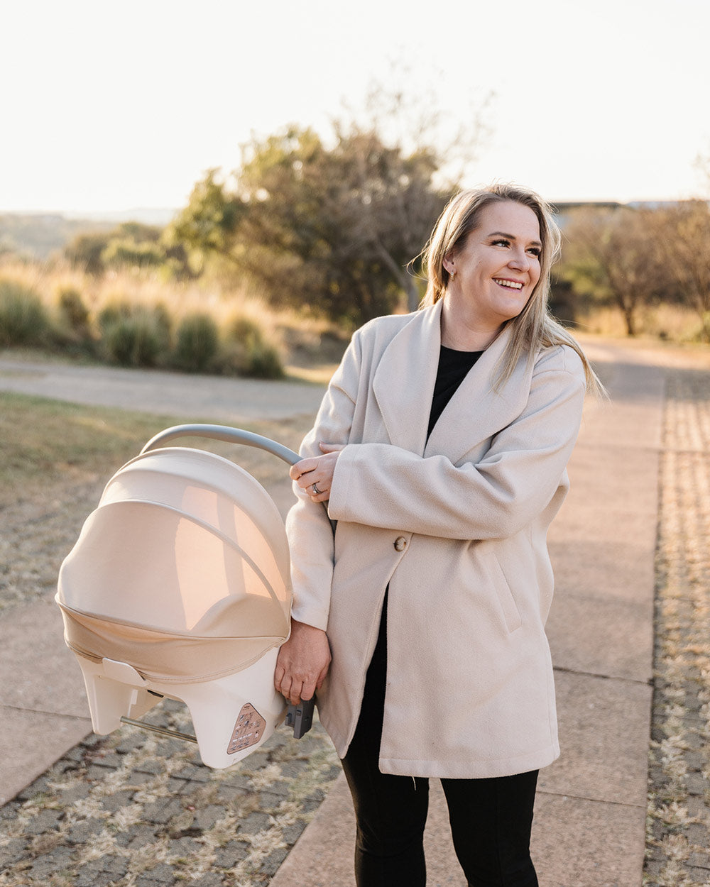 Woman smiling while carrying the BeBeBus Guard Car Seat by its handle during a walk outdoors.