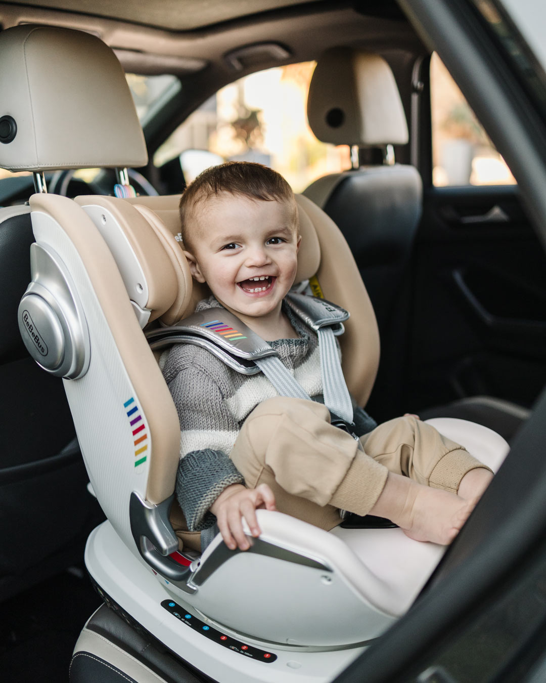 Smiling toddler sitting in BeBeBus Astron+ PRO Car Seat with secure 5-point harness inside a car.