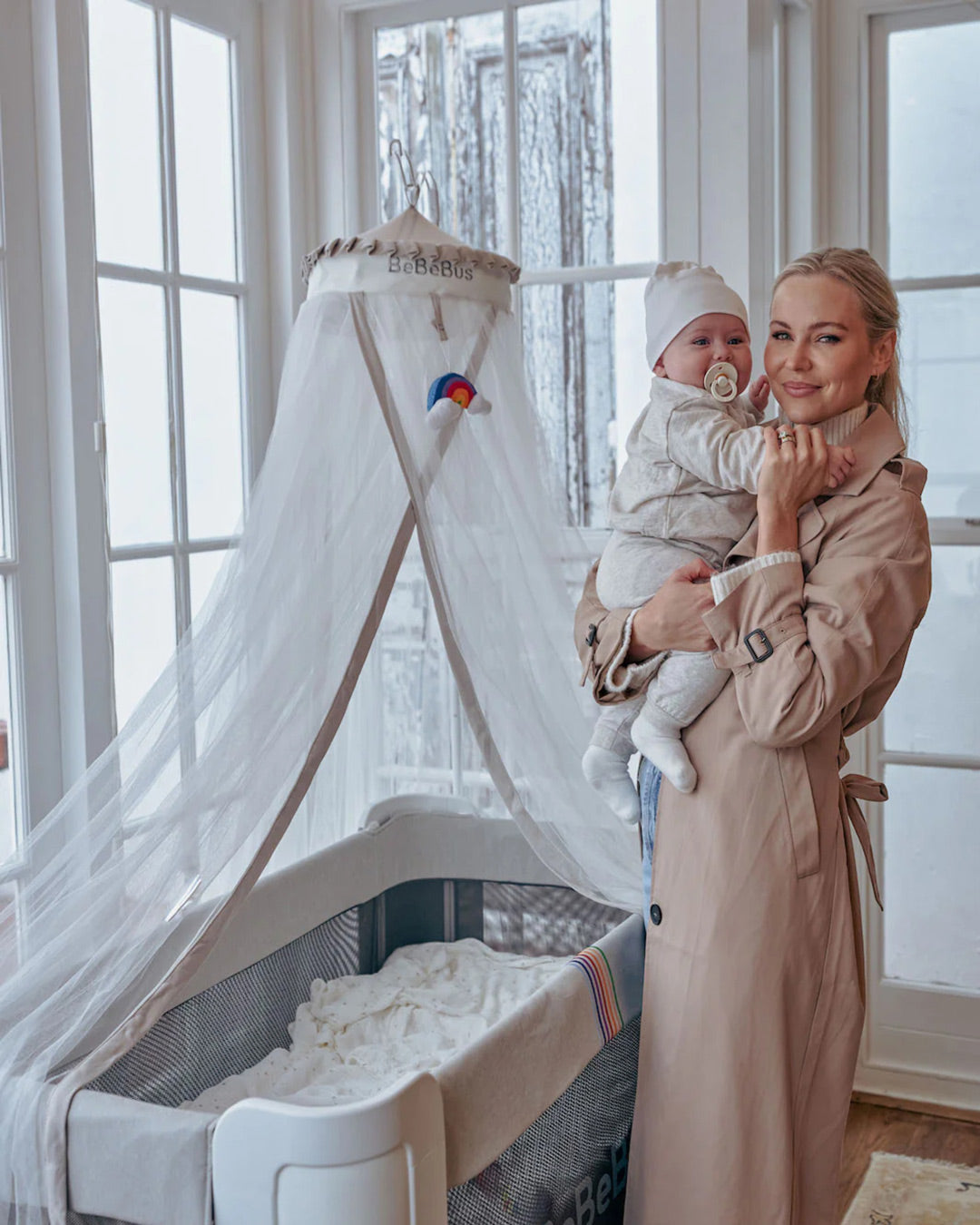 Mom holding baby beside BeBeBus crib fitted with a white mosquito net canopy, a functional nursery accessory and baby sleep essential for peaceful naps.