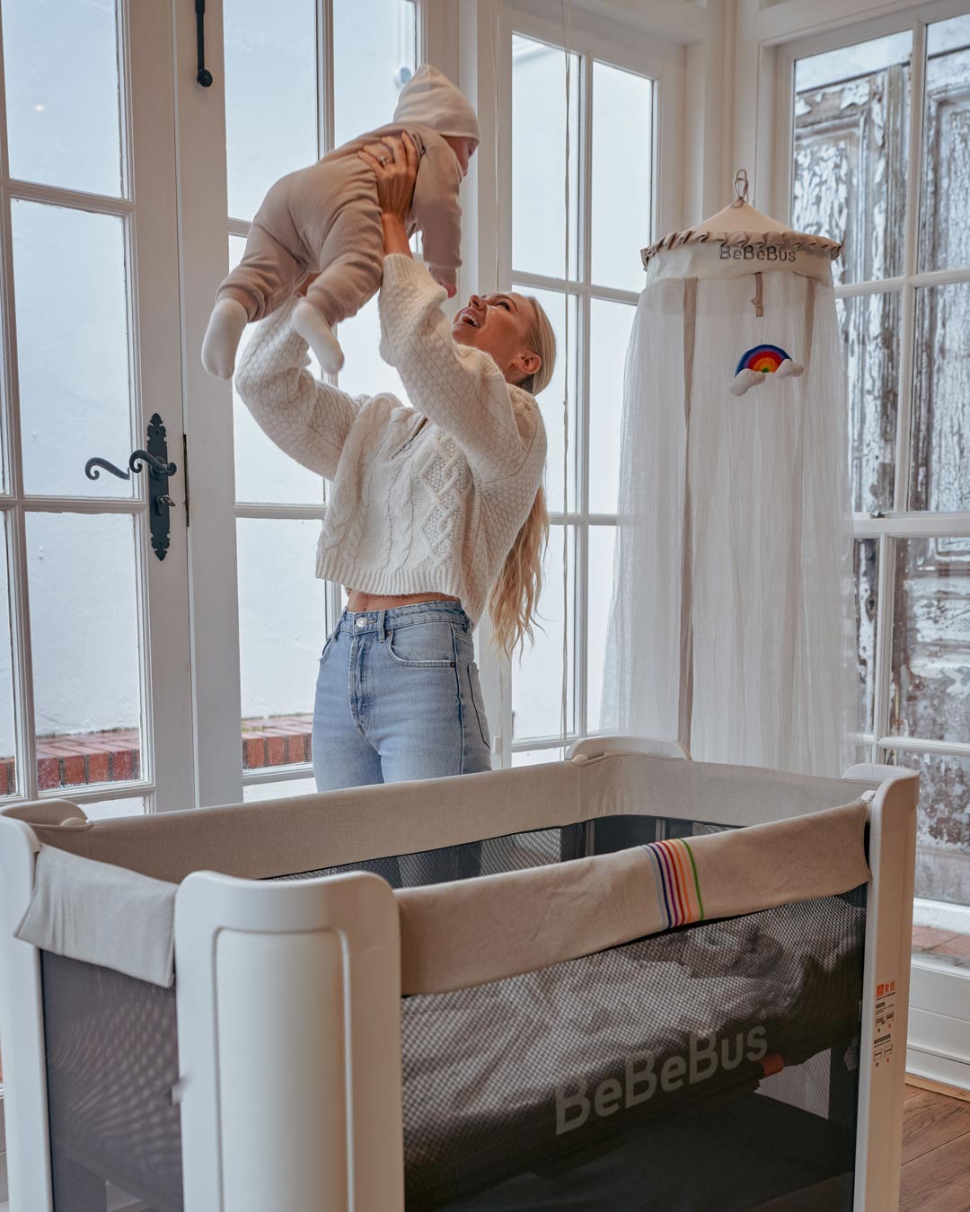 A happy mom lifts her baby in front of the BeBeBus crib with a freestanding mosquito net in a bright, airy room.