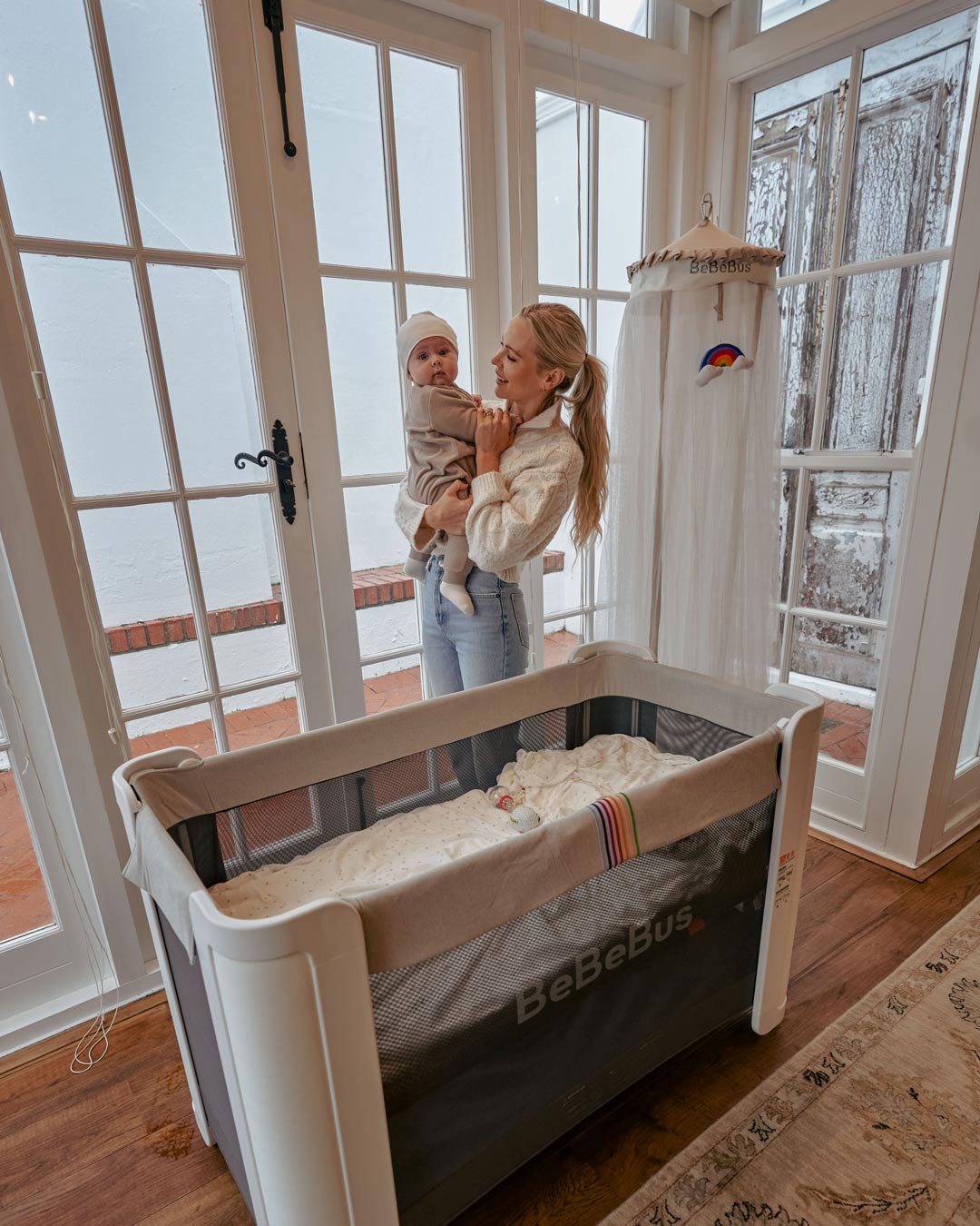 A mother holds her baby next to the BeBeBus crib and freestanding mosquito net, creating a peaceful nursery environment.
