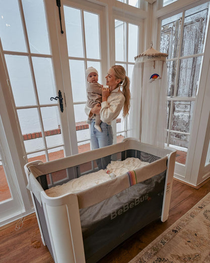 A mother holds her baby next to the BeBeBus crib and freestanding mosquito net, creating a peaceful nursery environment.